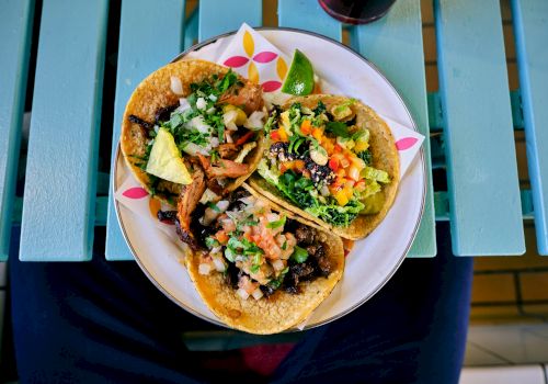 A plate with three colorful tacos filled with various ingredients like meat, vegetables, and avocado, placed on a blue wooden table.