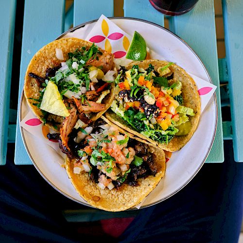 A plate with three colorful tacos filled with various ingredients like meat, vegetables, and avocado, placed on a blue wooden table.