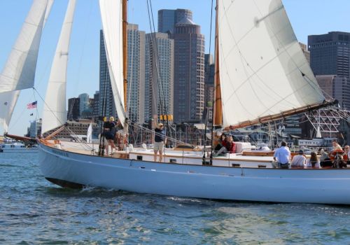 A sailboat with people onboard is sailing in the water, with a cityscape featuring tall buildings in the background.