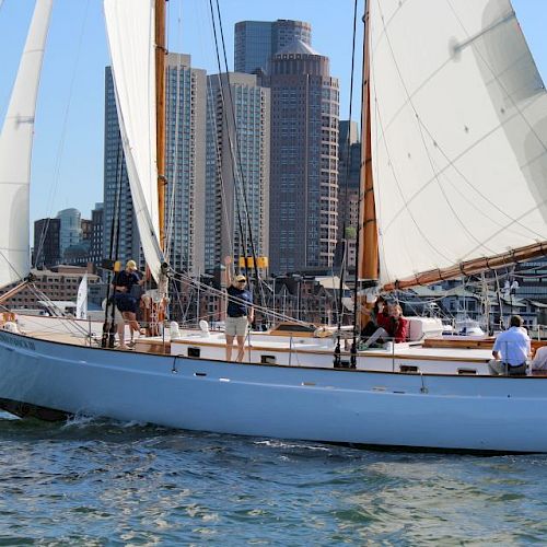 A sailboat with people onboard is sailing in the water, with a cityscape featuring tall buildings in the background.