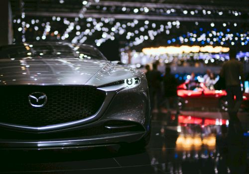 A sleek, silver Mazda car is displayed at an auto show, with bright lights and blurred people in the background.