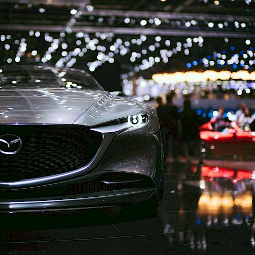 A sleek, silver Mazda car is displayed at an auto show, with bright lights and blurred people in the background.