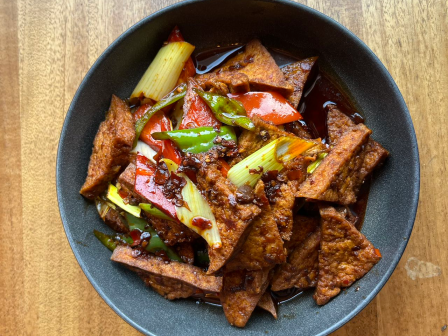 The image shows a dish consisting of stir-fried tofu with vegetables like bell peppers, scallions, and onions, served in a black bowl on a wooden surface.