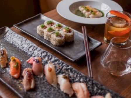 This image shows a dining table with an assortment of sushi, including nigiri and rolls, a pair of wooden chopsticks, and drinks.