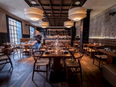 The image shows a cozy restaurant interior with wooden furniture, pendant lights, and set tables, featuring a blurred person moving in the background.