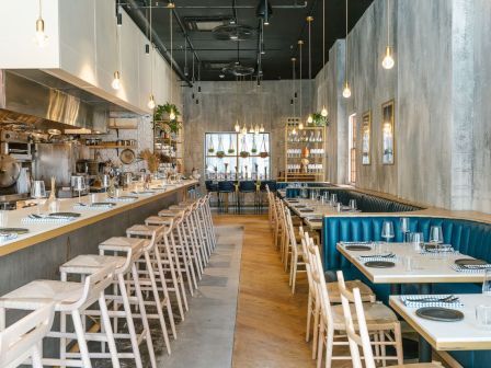 The image shows a modern, well-lit restaurant with a bar area, wooden chairs, blue booth seating, and tables set for dining.