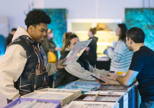 People browsing and chatting at a record store or market, with a focus on a person holding a vinyl record while another looks on interested.