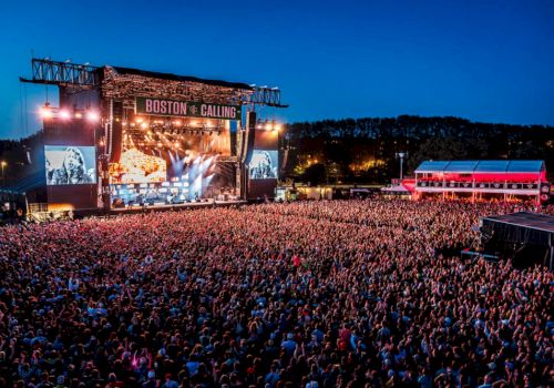 A large outdoor concert with a huge crowd and a stage lit up with colorful lights at night. The event is named 