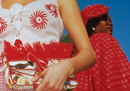 The image shows two people; one in a sleeveless top with red designs holding a patterned red purse, the other wearing a red checkered outfit and headscarf.