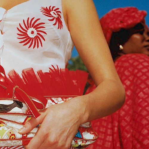 The image shows two people; one in a sleeveless top with red designs holding a patterned red purse, the other wearing a red checkered outfit and headscarf.