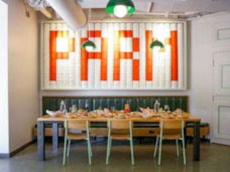 The image shows a dining area with a long wooden table and chairs. The wall has a large, brightly lit sign that reads 