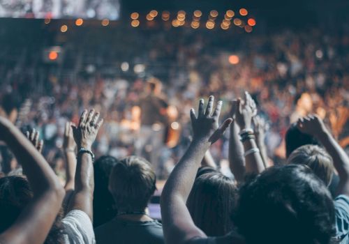 People are raising their hands in a crowded, lively event, likely a concert or a large gathering, with vibrant lights and a blurred background.