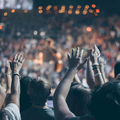 A lively crowd enjoys a concert or event, with hands raised in the air, while blurred lights and people create a vibrant atmosphere in the background.