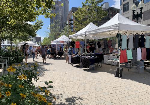 An outdoor market with white tents selling clothes and various items, people walking around, and buildings in the background, on a sunny day.