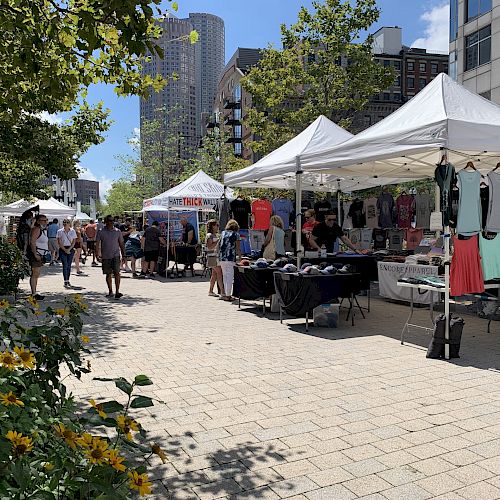 An outdoor market with white tents selling clothes and various items, people walking around, and buildings in the background, on a sunny day.