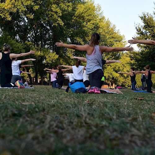 A group of people practicing yoga outdoors, standing on mats with arms outstretched, surrounded by trees and grass.