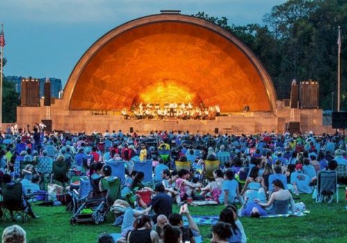 People are seated on the lawn, watching a live performance on an outdoor stage with an orange-lit arch, during evening time.