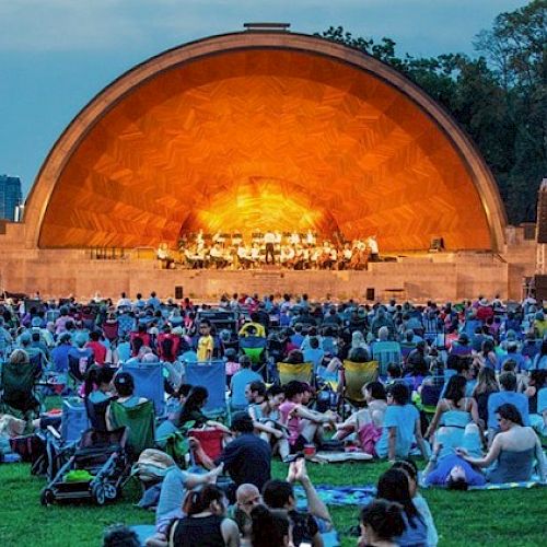 People are seated on the lawn, watching a live performance on an outdoor stage with an orange-lit arch, during evening time.