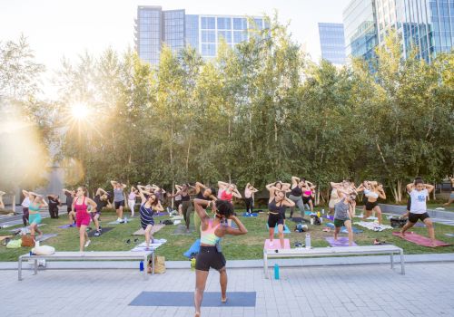 A group of people is participating in an outdoor fitness class on yoga mats in a park with a cityscape in the background, at sunset.