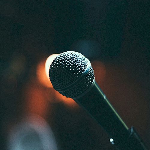 A close-up of a microphone on a stand, with a dimly lit background featuring blurred lights.