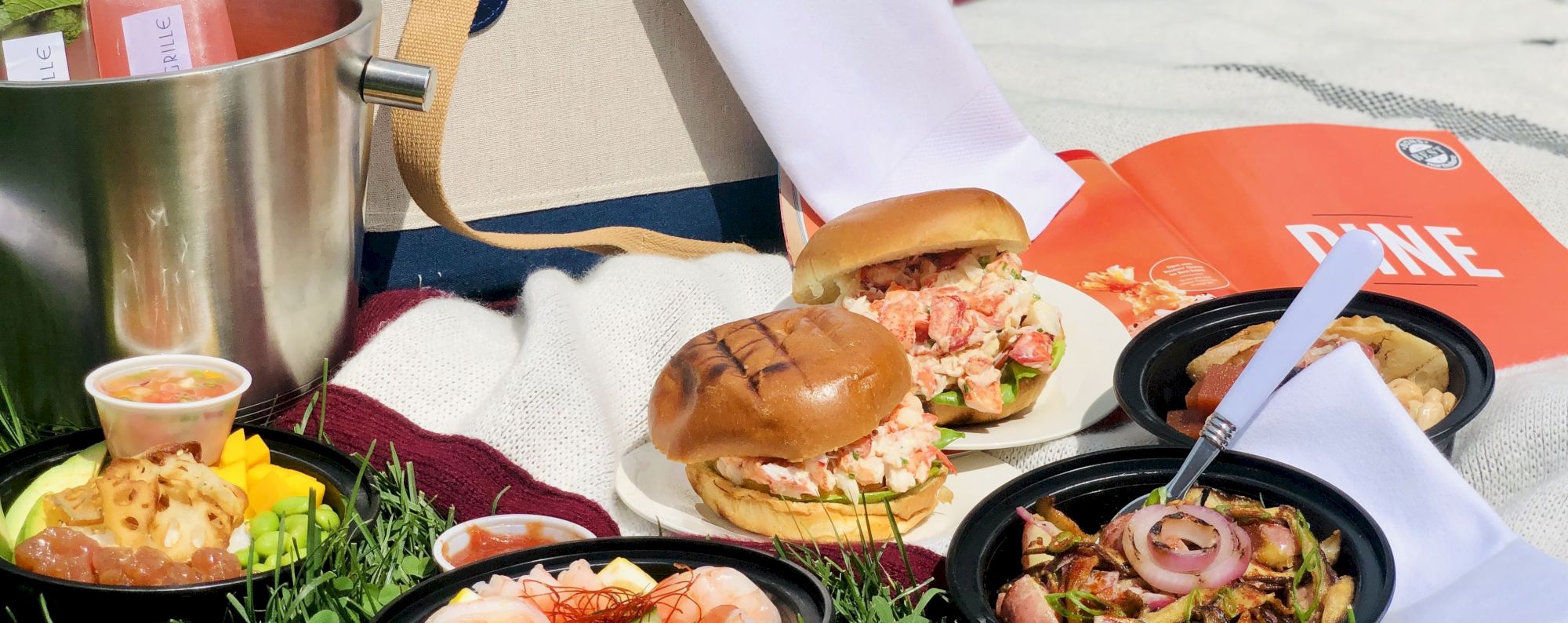 Picnic setup with sandwiches, salads, drinks, and a basket on a sunny day.