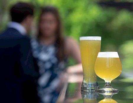 Two glasses of beer in focus with blurry people in the background.