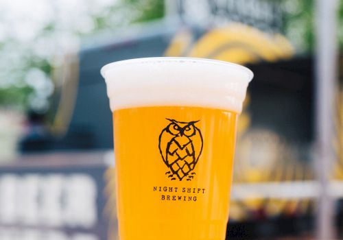 A plastic cup filled with a frothy beer from “Night Shift Brewing” is placed on a wooden surface, with a blurred background of outdoor scene.