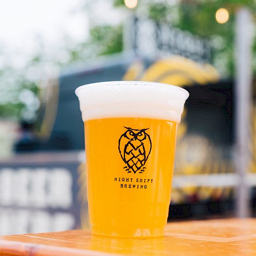 A plastic cup filled with a frothy beer from “Night Shift Brewing” is placed on a wooden surface, with a blurred background of outdoor scene.