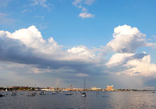 A serene waterside view featuring numerous boats, a distant shoreline with buildings, and a sky filled with large, fluffy clouds ending the sentence.