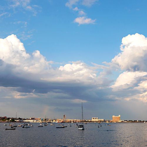 A serene waterside view featuring numerous boats, a distant shoreline with buildings, and a sky filled with large, fluffy clouds ending the sentence.