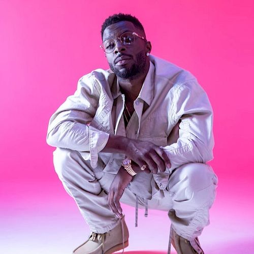 A man squats, wearing white clothes, against a pink backdrop.