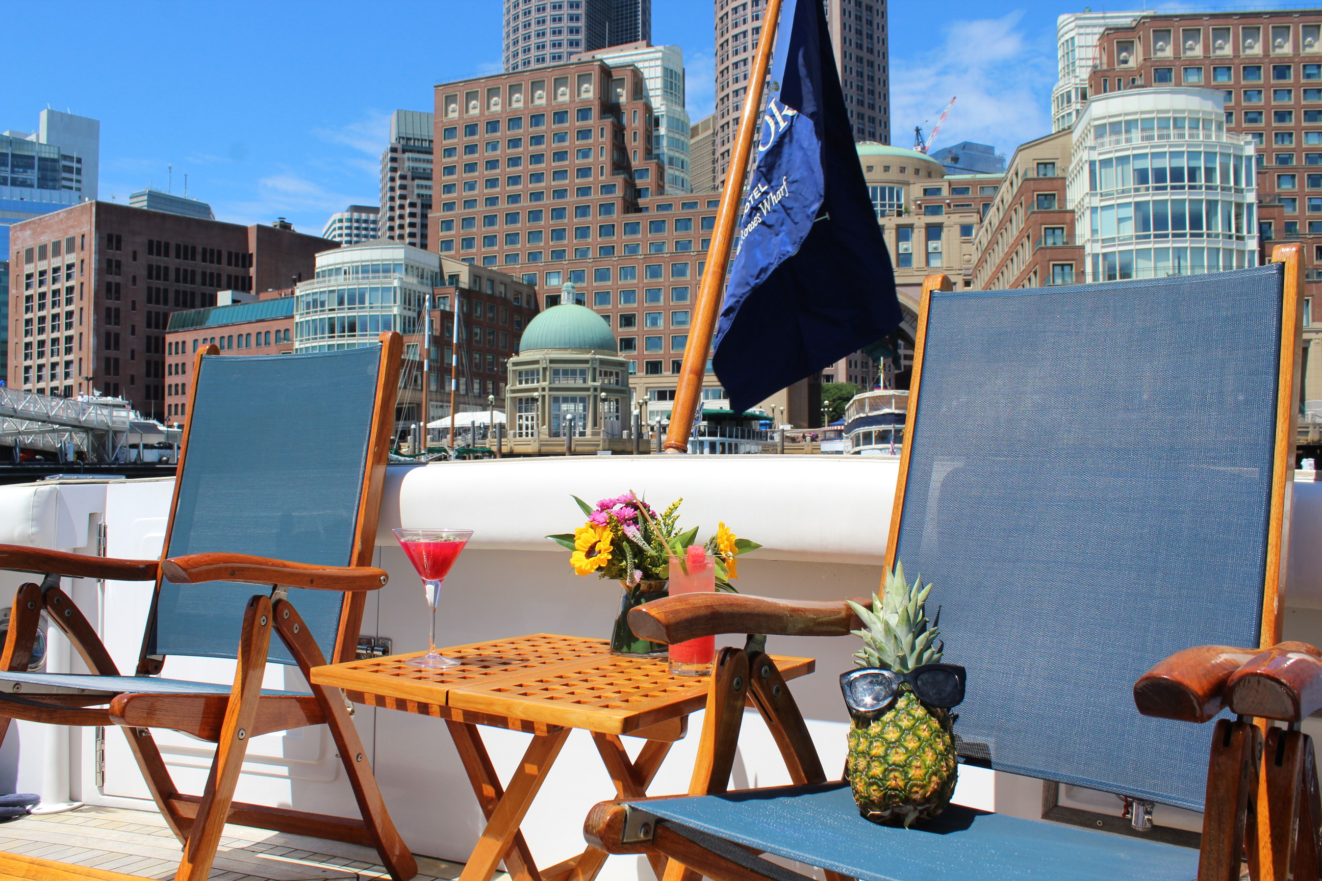 Two lounge chairs, a small table with cocktails and flowers, and a pineapple wearing sunglasses on a boat, with city buildings in the background.