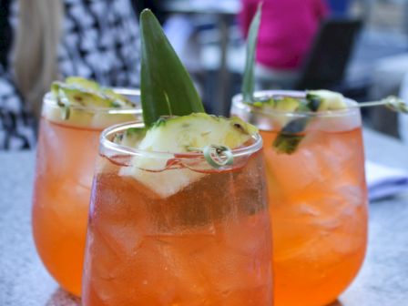 Three glasses of orange cocktails with ice, garnished with pineapple slices and leaves, set on a table outdoors.