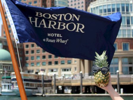 A hand holds a pineapple with sunglasses in front of a Boston Harbor Hotel flag and buildings in the background, creating a fun scene.