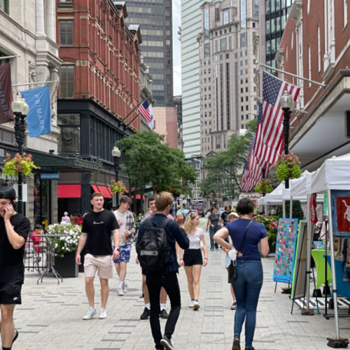 People are walking in a bustling urban street filled with shops, including Macy's, and outdoor vendors, with American flags displayed.