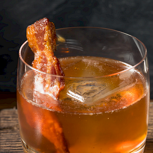 A cocktail garnished with a piece of crispy bacon, served in a glass with an ice cube, placed on a wooden surface.