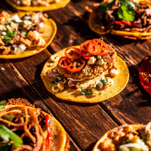 An assortment of colorful tacos with various toppings, including vegetables, meats, and garnishes, arranged on a rustic wooden surface.