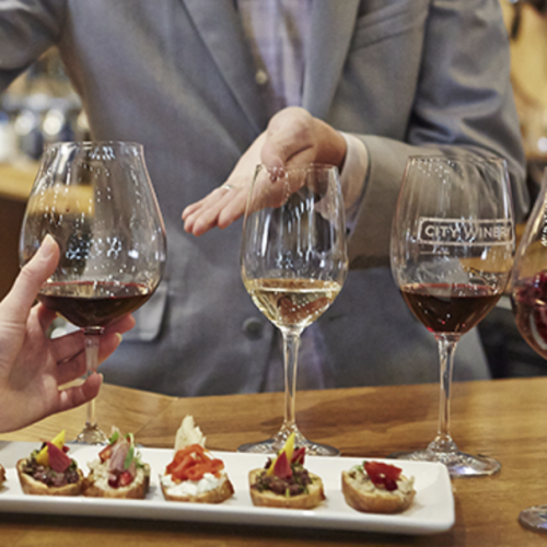 A group of people participating in a wine tasting, holding glasses, with several appetizers on a plate in front of them, ending the sentence.