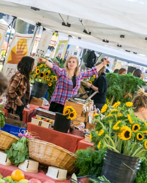 People are shopping at an outdoor market with stalls selling flowers, produce, and other goods under white tents.