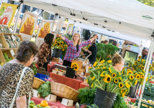 People are shopping at an outdoor market with stalls selling flowers, produce, and other goods under white tents.