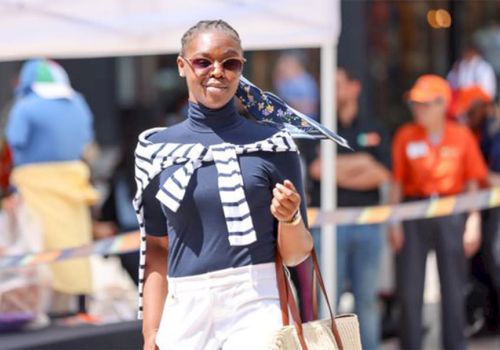 A person wearing sunglasses, striped sweater, and navy top walks confidently outside, carrying a tote bag and surrounded by blurred background figures.