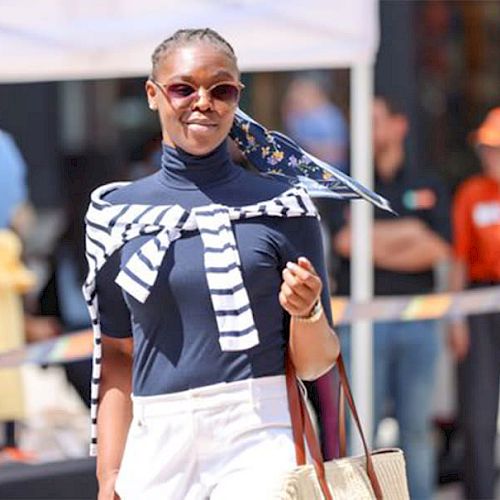 A person wearing sunglasses, striped sweater, and navy top walks confidently outside, carrying a tote bag and surrounded by blurred background figures.