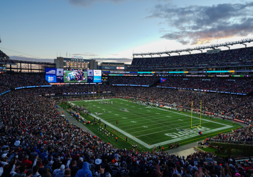 The image shows a packed football stadium during a game, with players on the field, large screens displaying information, and a crowd in the stands.