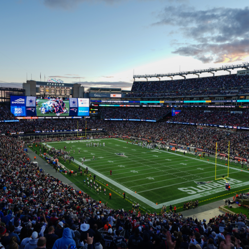 The image shows a packed football stadium during a game, with players on the field, large screens displaying information, and a crowd in the stands.