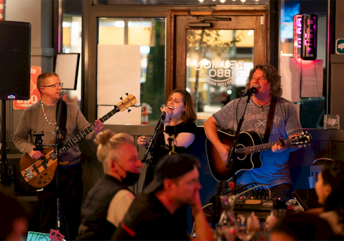 Three musicians perform on a small stage in a lively restaurant or bar, entertaining a crowd of seated patrons.