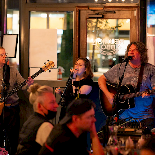 Three musicians perform on a small stage in a lively restaurant or bar, entertaining a crowd of seated patrons.