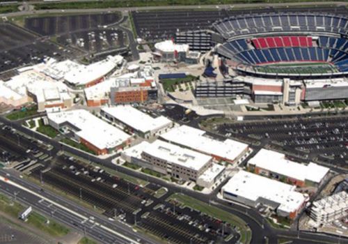 An aerial view of a large event stadium surrounded by numerous buildings and extensive parking lots, with major roads visible in the foreground.