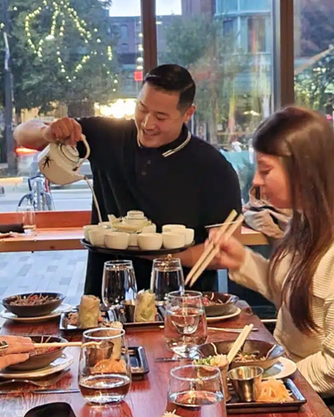 A server is pouring tea for a group of people dining at a restaurant. Various dishes and drinks are on the table, and the group is engaged and smiling.