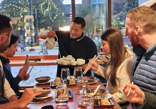 A server is pouring tea for a group of people dining at a restaurant. Various dishes and drinks are on the table, and the group is engaged and smiling.