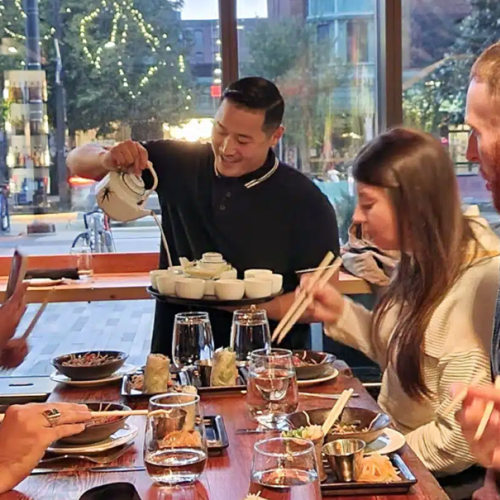 A server is pouring tea for a group of people dining at a restaurant. Various dishes and drinks are on the table, and the group is engaged and smiling.